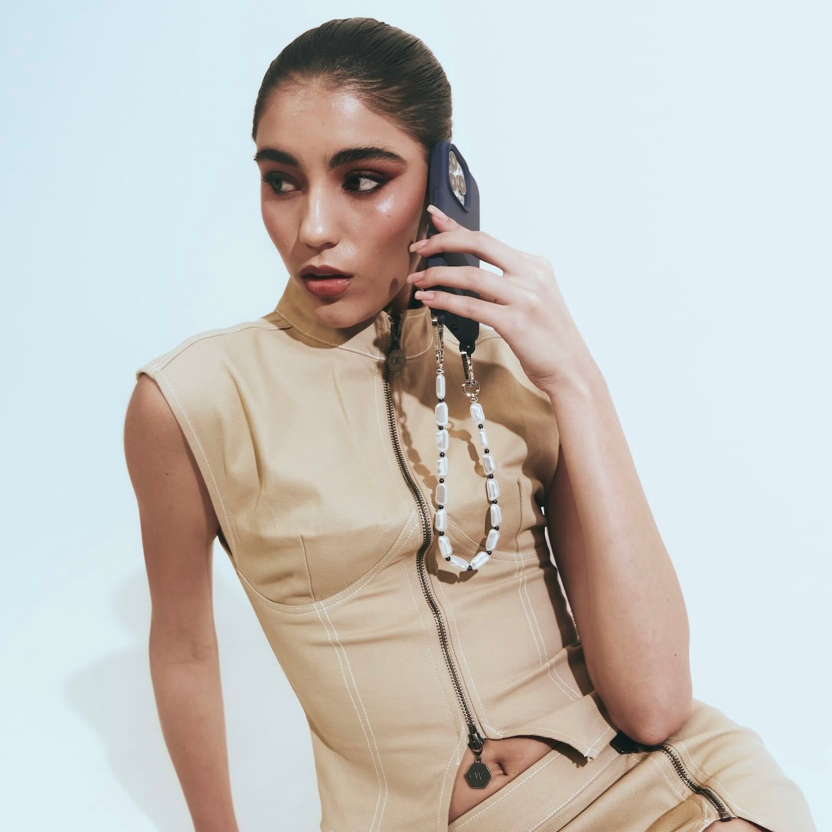 Woman in beige outfit with a phone chain, holding a phone to her ear against a light background